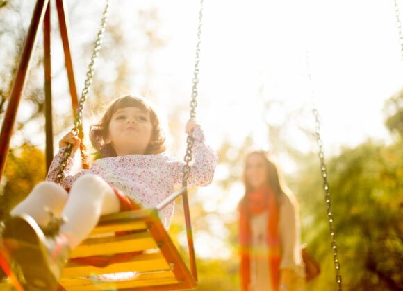 A young girl swings in a sunlit park while an adult watches nearby.