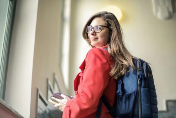 A woman with long hair and glasses wearing a red coat and carrying a backpack looks back while holding a book.