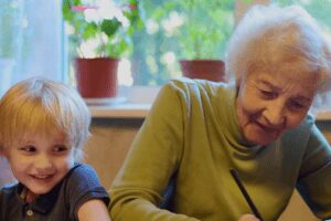 An elderly woman and a young child smiling together while drawing at a table.