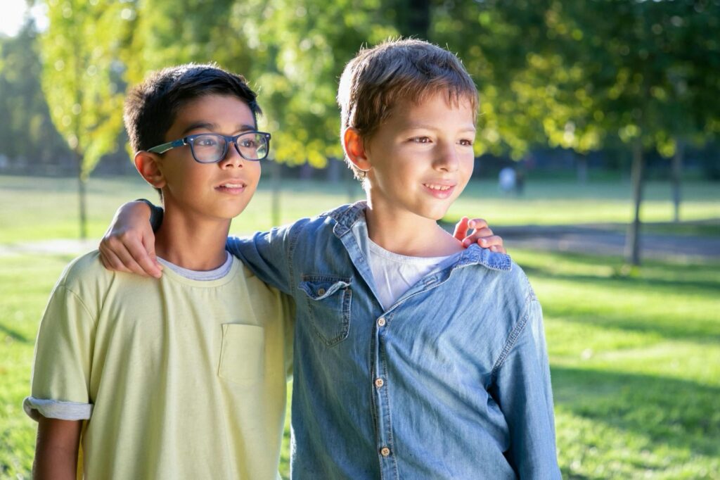 Two boys stand together in a park, smiling and enjoying the outdoors.